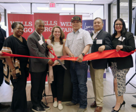 Several people stand behind a ribbon as the person near the center cuts the ribbon.