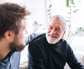 An older adult sits on the couch smiling. A younger adult sits next to him with his arm around him.