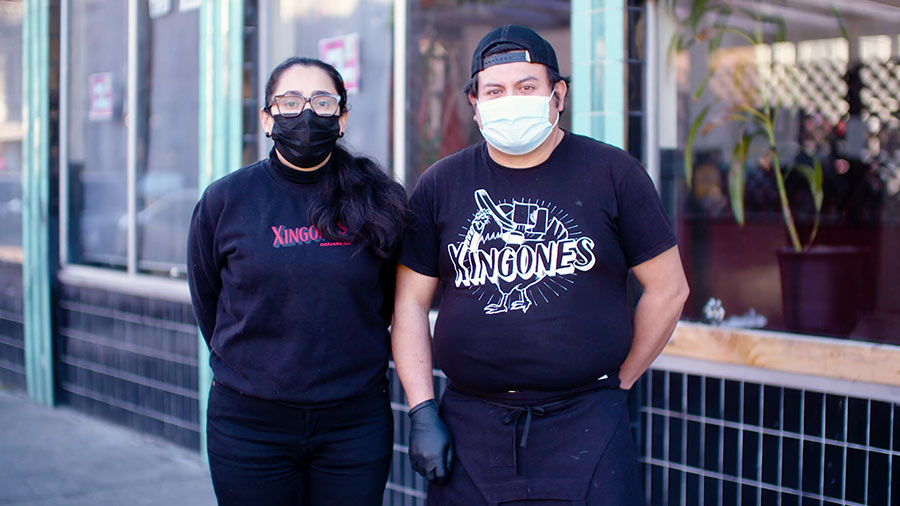 Two people with face masks on stand in front of a restaurant.