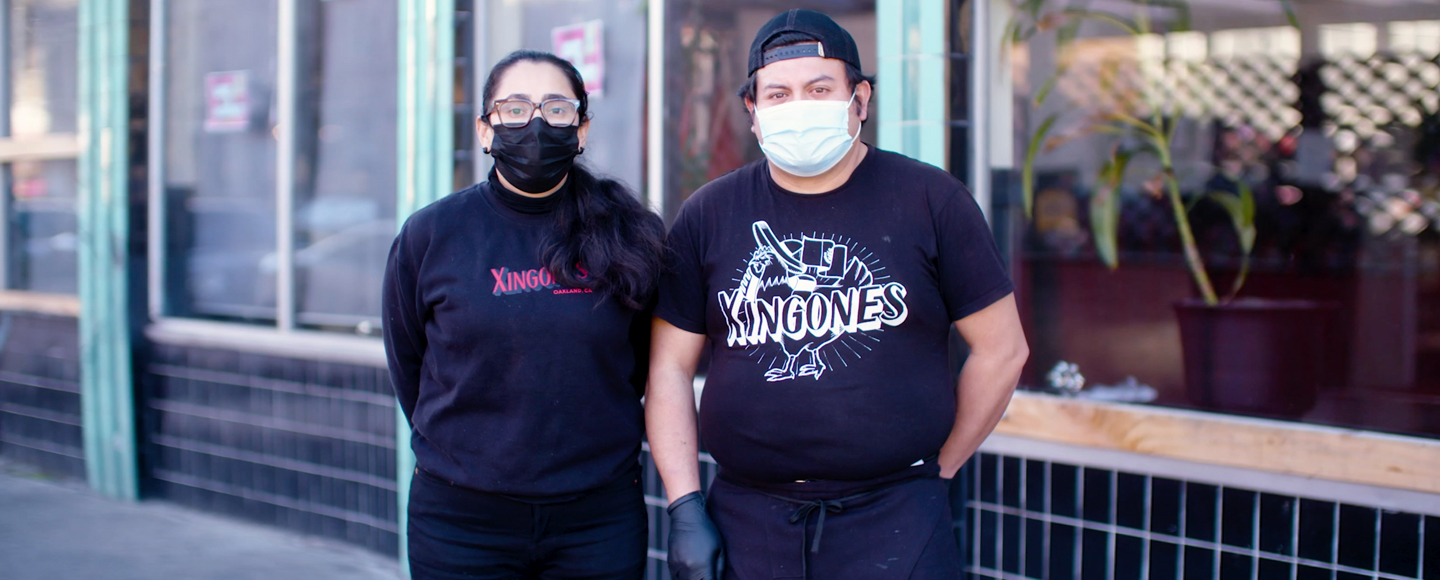 Two people with face masks on stand in front of a restaurant.