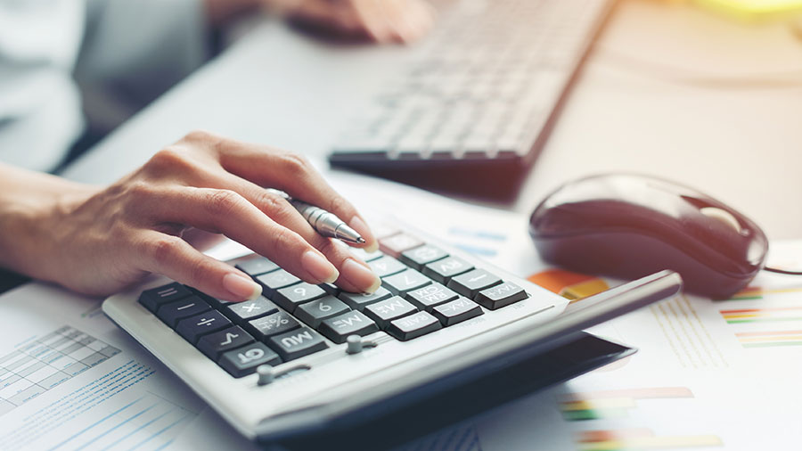 La imagen muestra una mano, con un bolígrafo entre dos dedos, que toca las teclas de una calculadora. Un teclado de computadora y un mouse aparecen en el fondo.