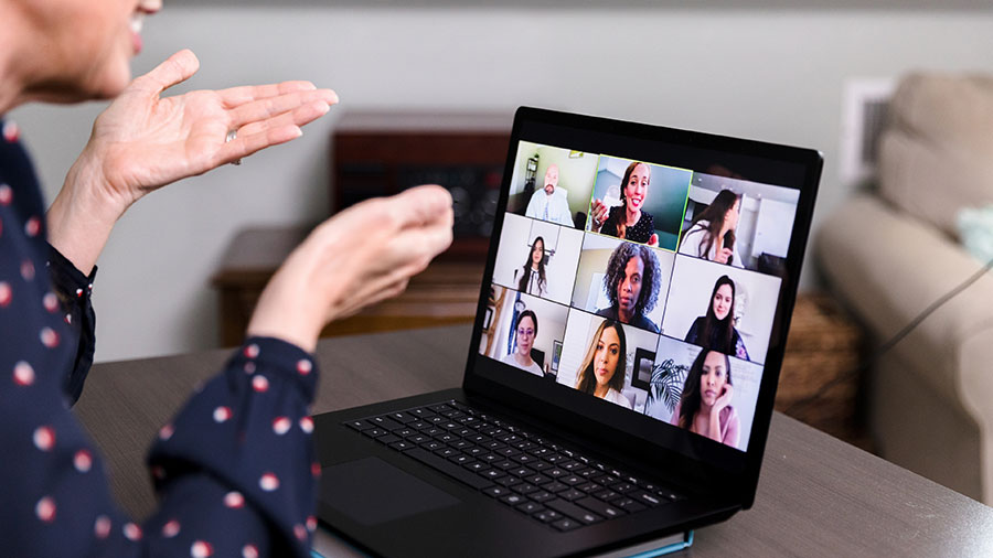 A woman gestures with her hands as she participates in a video conference call on her laptop with three rows of coworkers showing on the screen.