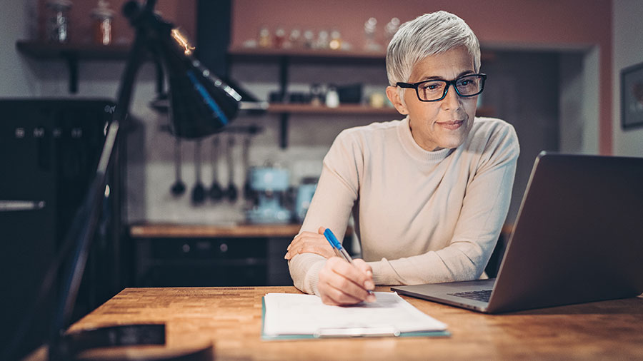 Una mujer con gafas se sienta en su escritorio y mira la pantalla de su computadora. Se prepara para anotar algo en un papel con el bolígrafo que sostiene en la mano derecha.