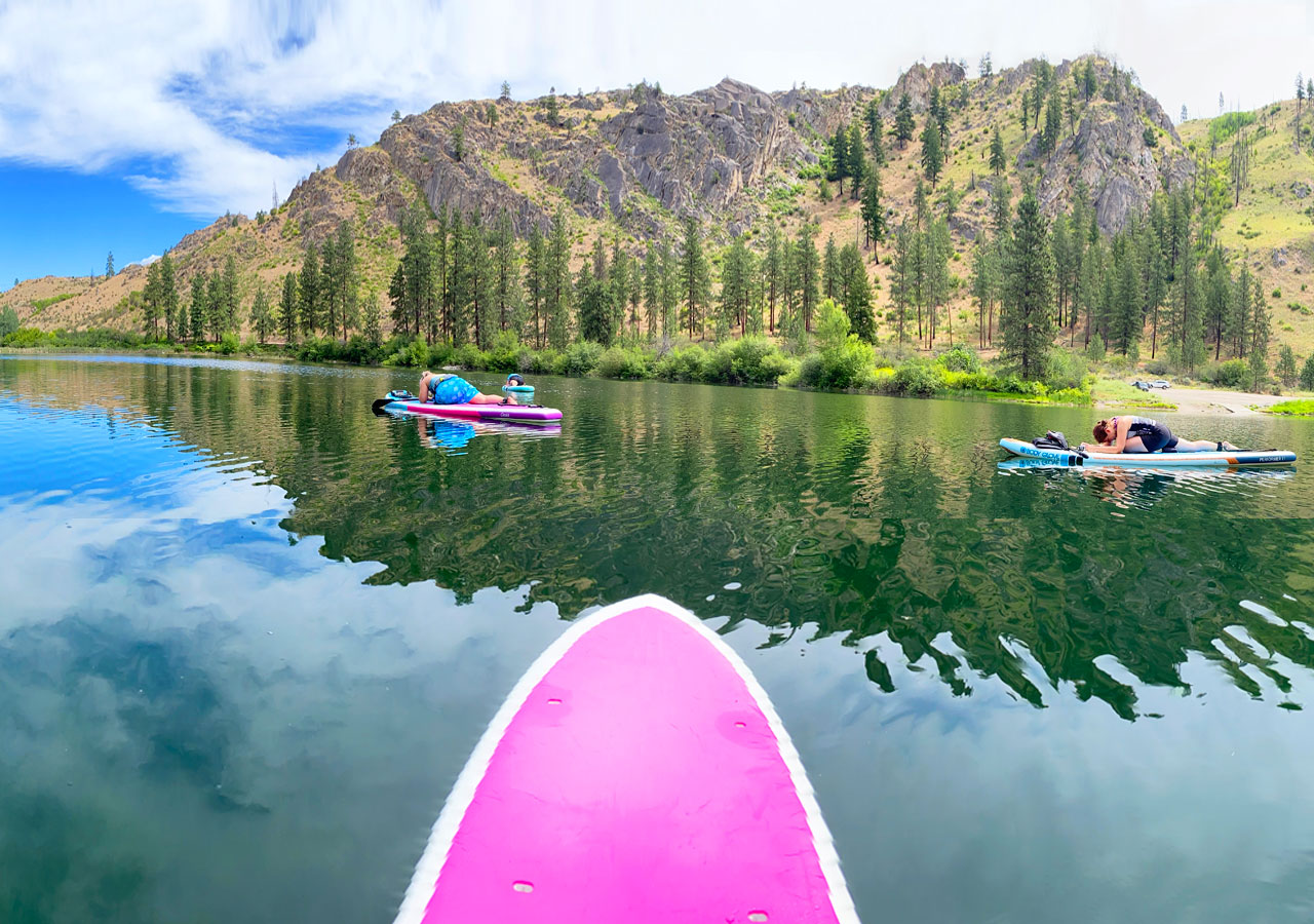  The front of a stand-up paddleboard is shown on the water and facing two people on other stand-up paddleboards. The body of water is in front of trees and a mountain.