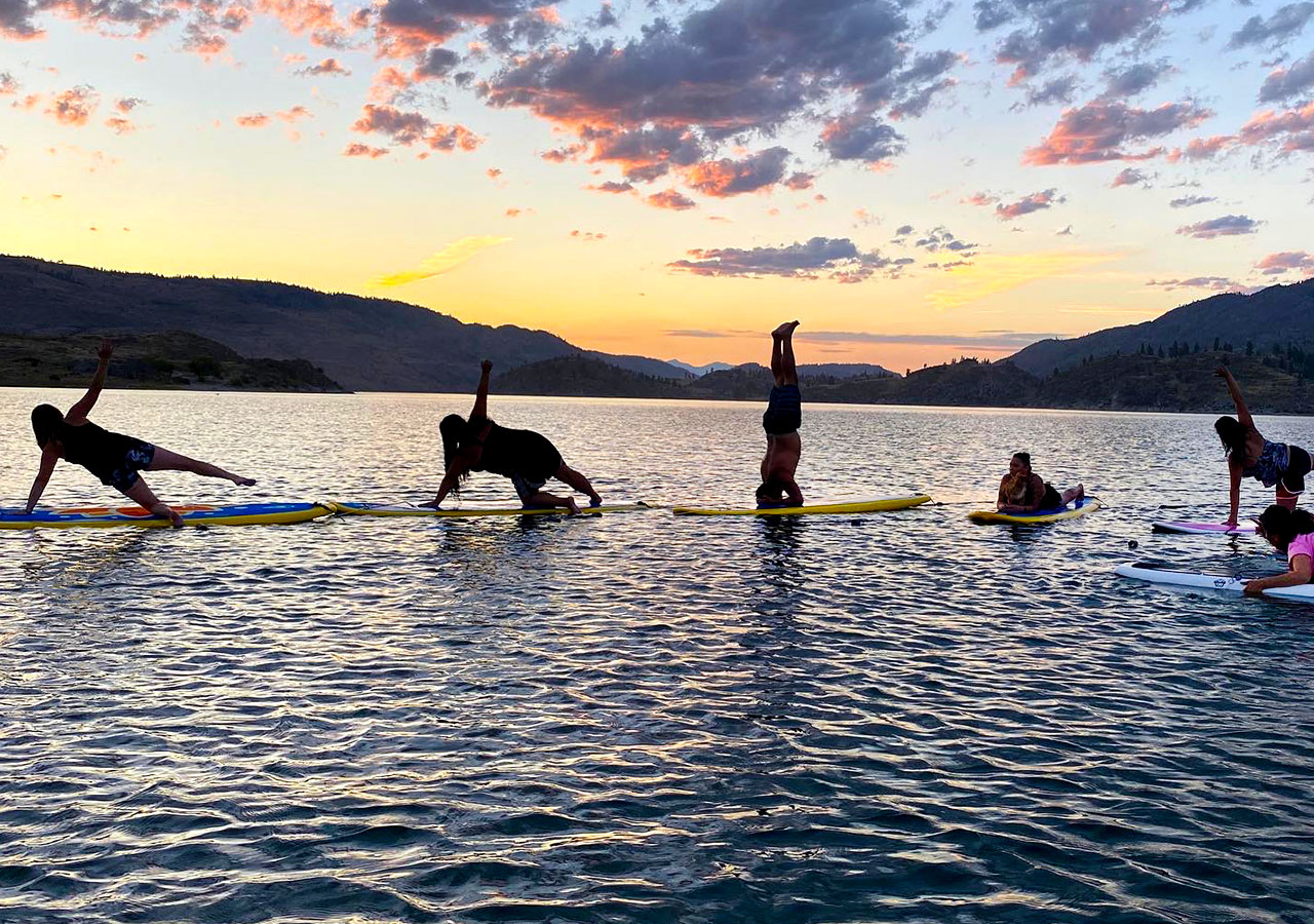  An image shows five people in various yoga poses while balancing on stand-up paddleboards on water. Behind them are mountains and a sunset.