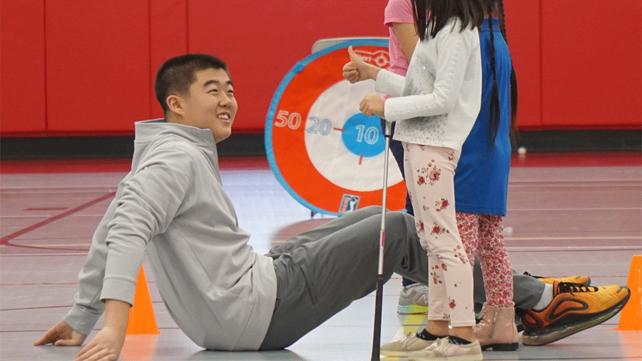 First Tee Greater Trenton's Raymond Jin looks up from a gym floor to a child holding a golf club and giving him the thumbs-up sign near a target with orange, blue and white circles and numbers.