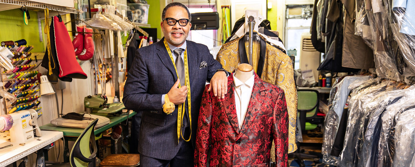 Eddie Lofton de pie en su tienda, JC Lofton Tailors, junto a un maniquí con traje y un estante de prendas en envoltorios de plástico.