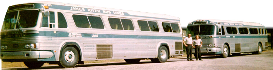 Two large gray buses with two men standing in front of them, hands in pockets.