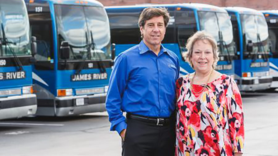 Man in blue shirt and woman in bright patterned blouse stand in front of blue buses labeled James River.
