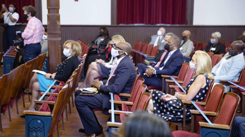 A crowd sits in an auditorium and listens to a speaker on stage.