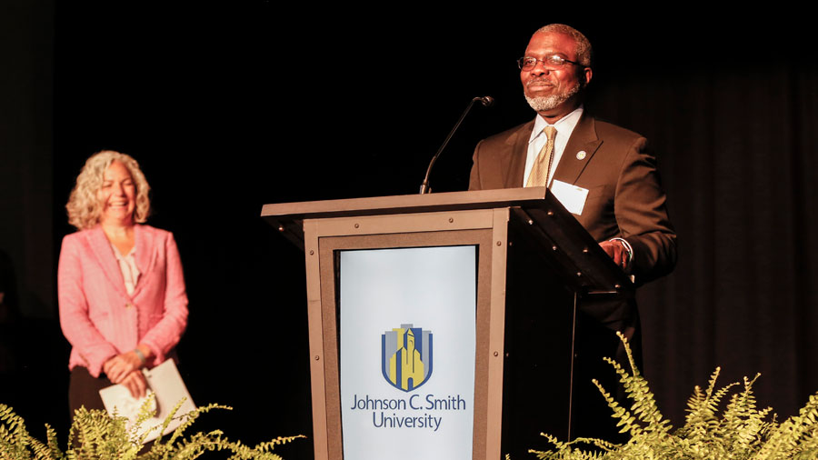 Johnson C. Smith University President Clarence Armbrister speaks at a podium with the school's logo on the front of it while Laura Clark from the United Way of Central Carolinas stands to the left holding notes and waiting for her turn to speak.