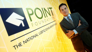Jorge Valencia, executive director and CEO of Point Foundation, stands in a suit with his arms crossed in front of a yellow banner that says Point Foundation, The National LGBTQ Scholarship Fund.