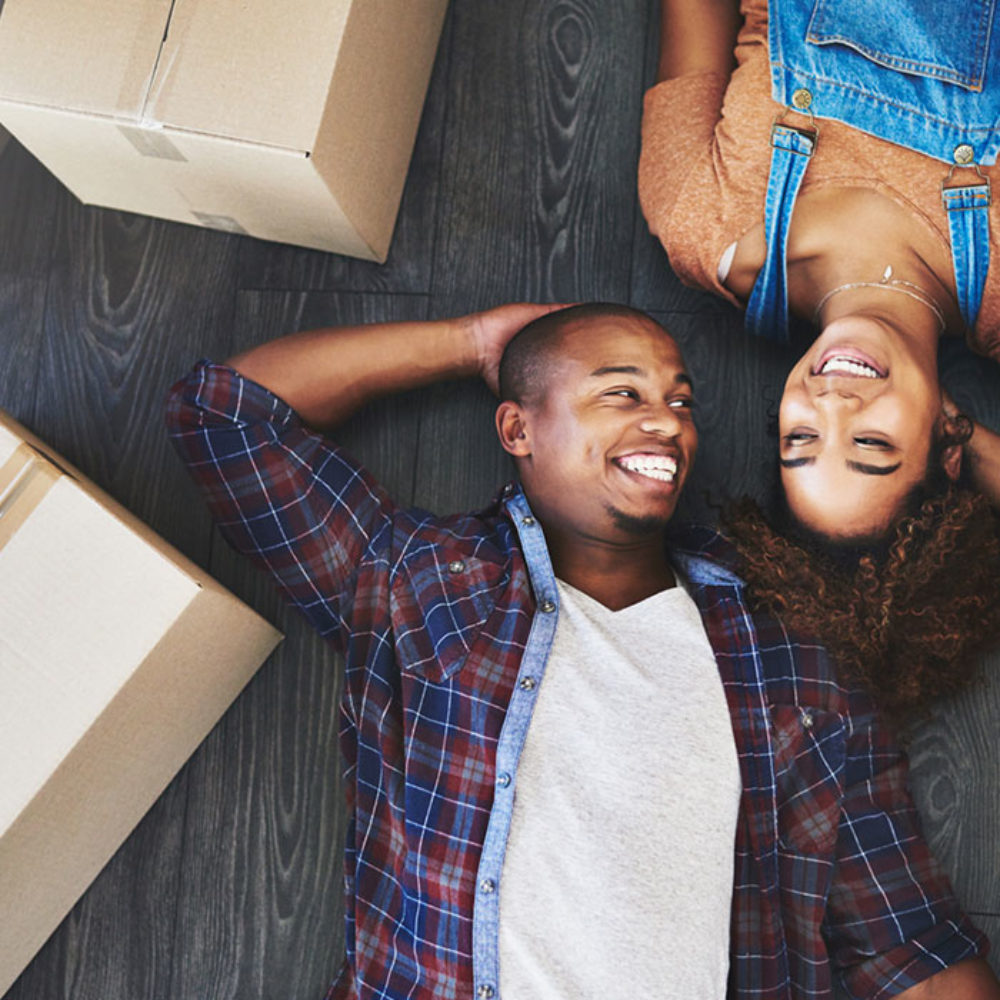 A man and a woman lie smiling on the floor surrounding by moving boxes.