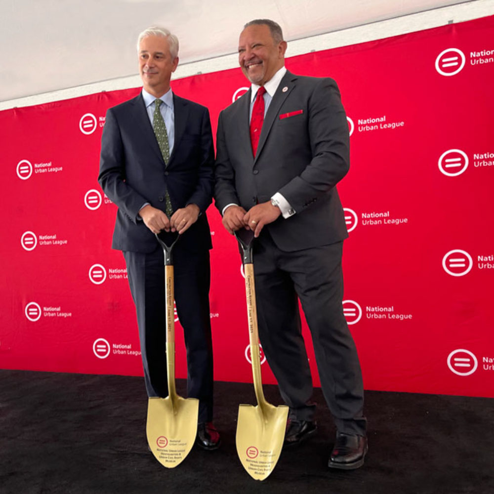 Charlie Scharf and Marc Morial stand, holding shovels, in front of a red banner that says National Urban League.