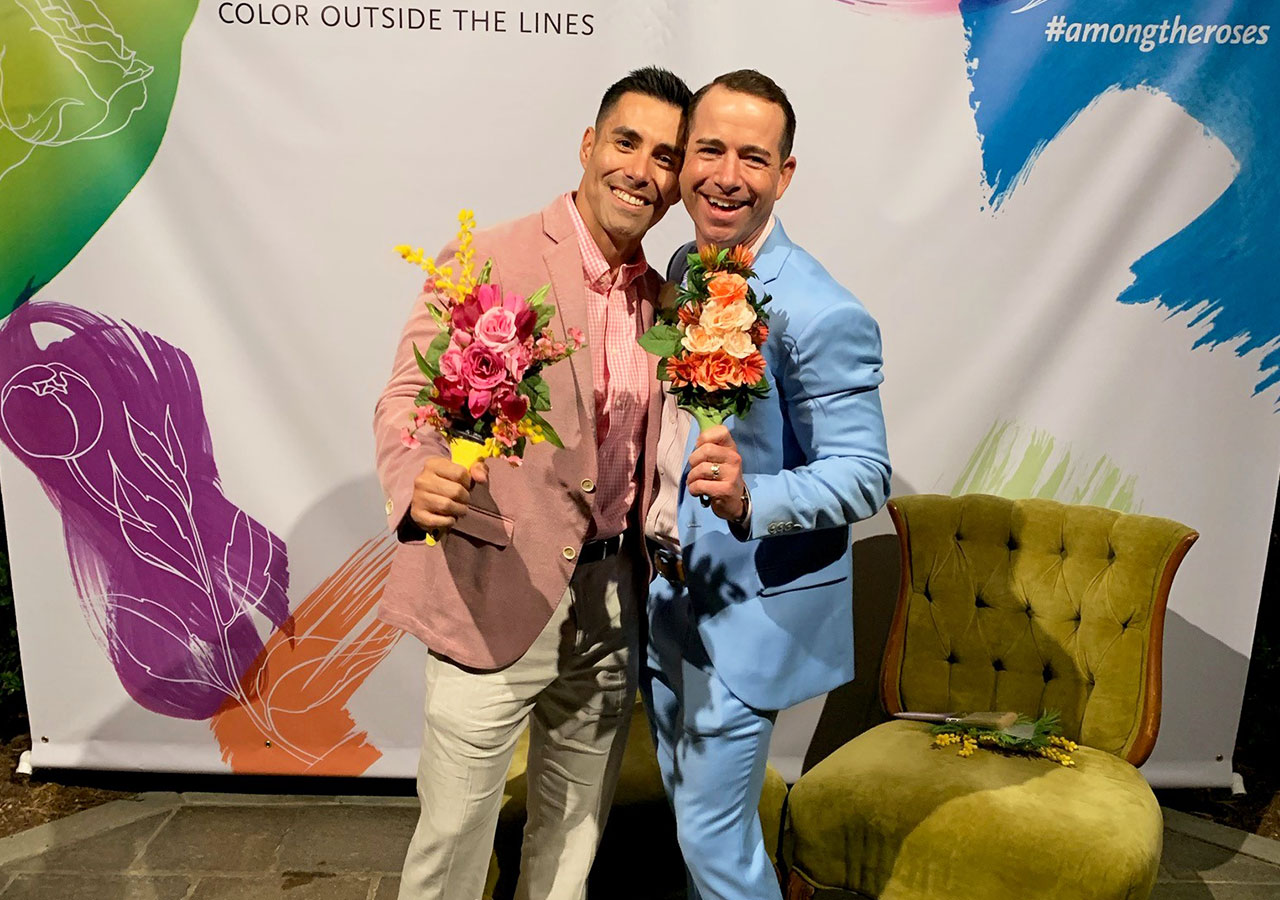  two men in pink and blue blazers hold small bouquets of flowers in front of a multicolored backdrop and gold armchair