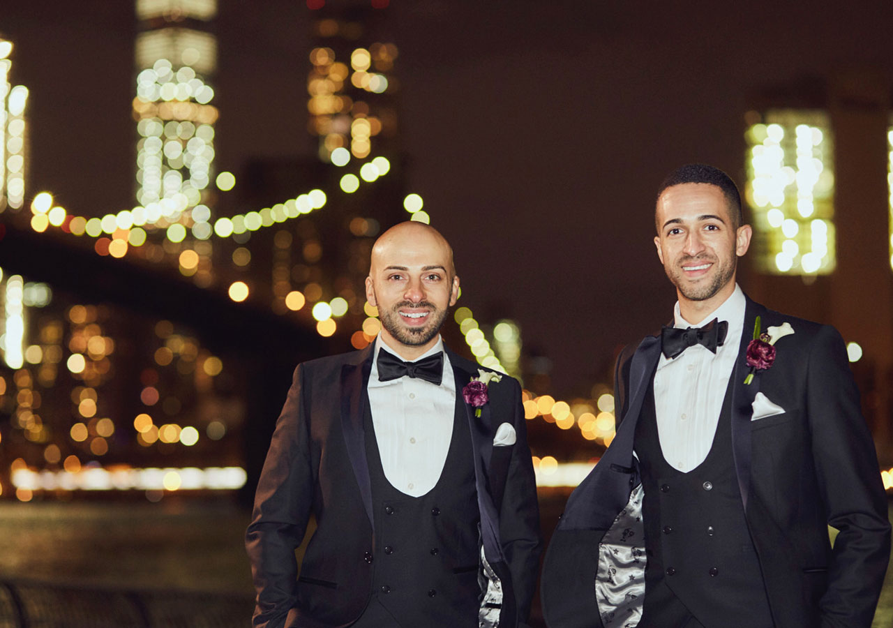  Two men stand before a skyline at night in dark tuxedo suits.