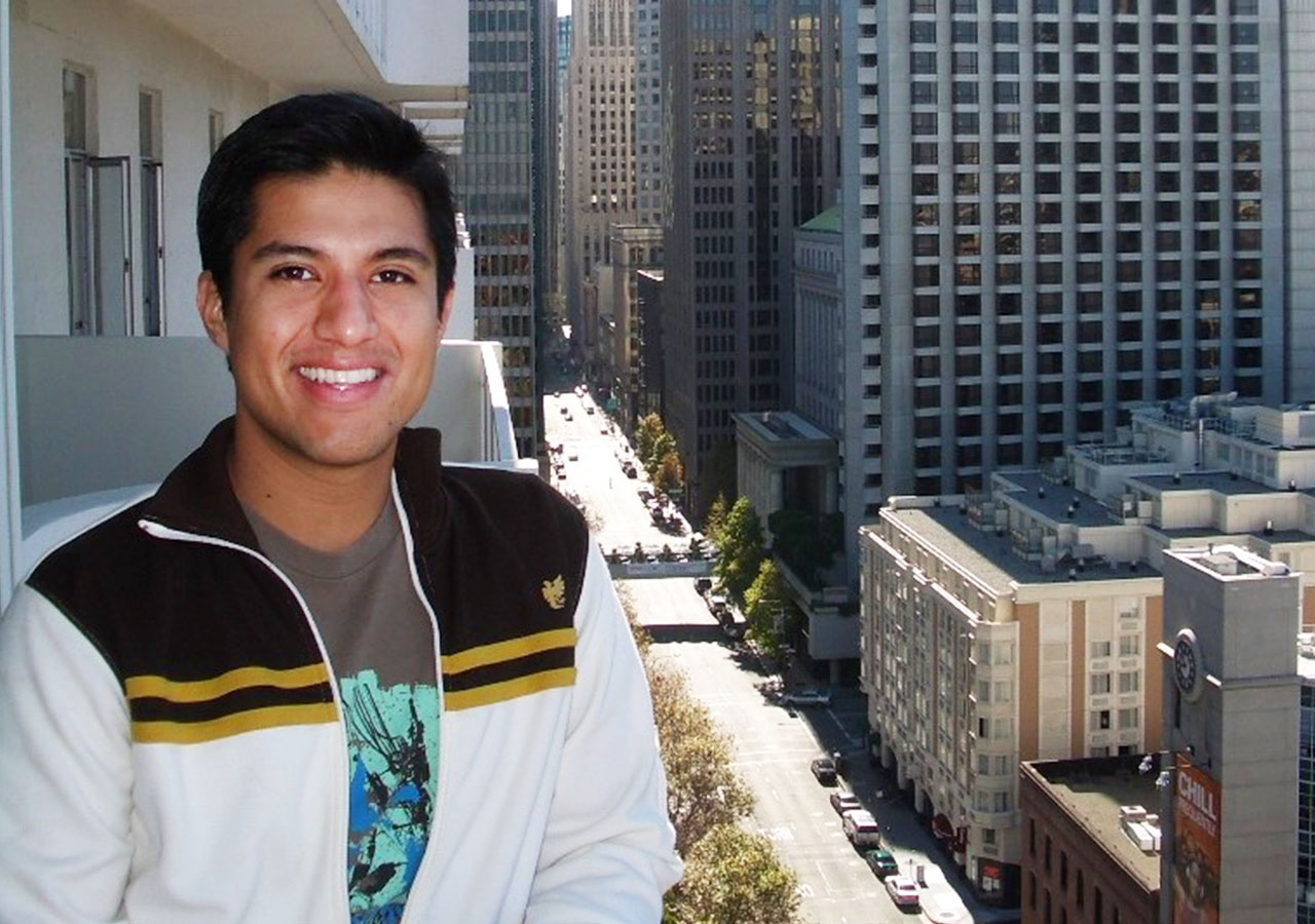  A younger Jason Vasquez is wearing a white jacket with dark stripes, and is standing on the balcony of a building in downtown San Francisco. The skyline and street are seen behind him.