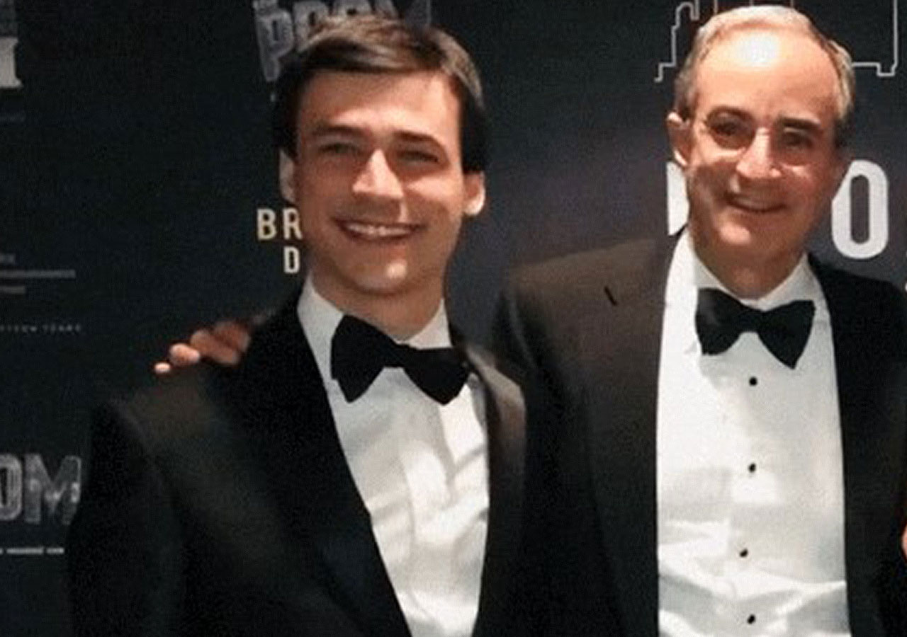  John Weiss, right, and his son both wear black bow ties and dark tuxedoes and stand before a black printed gala backdrop.