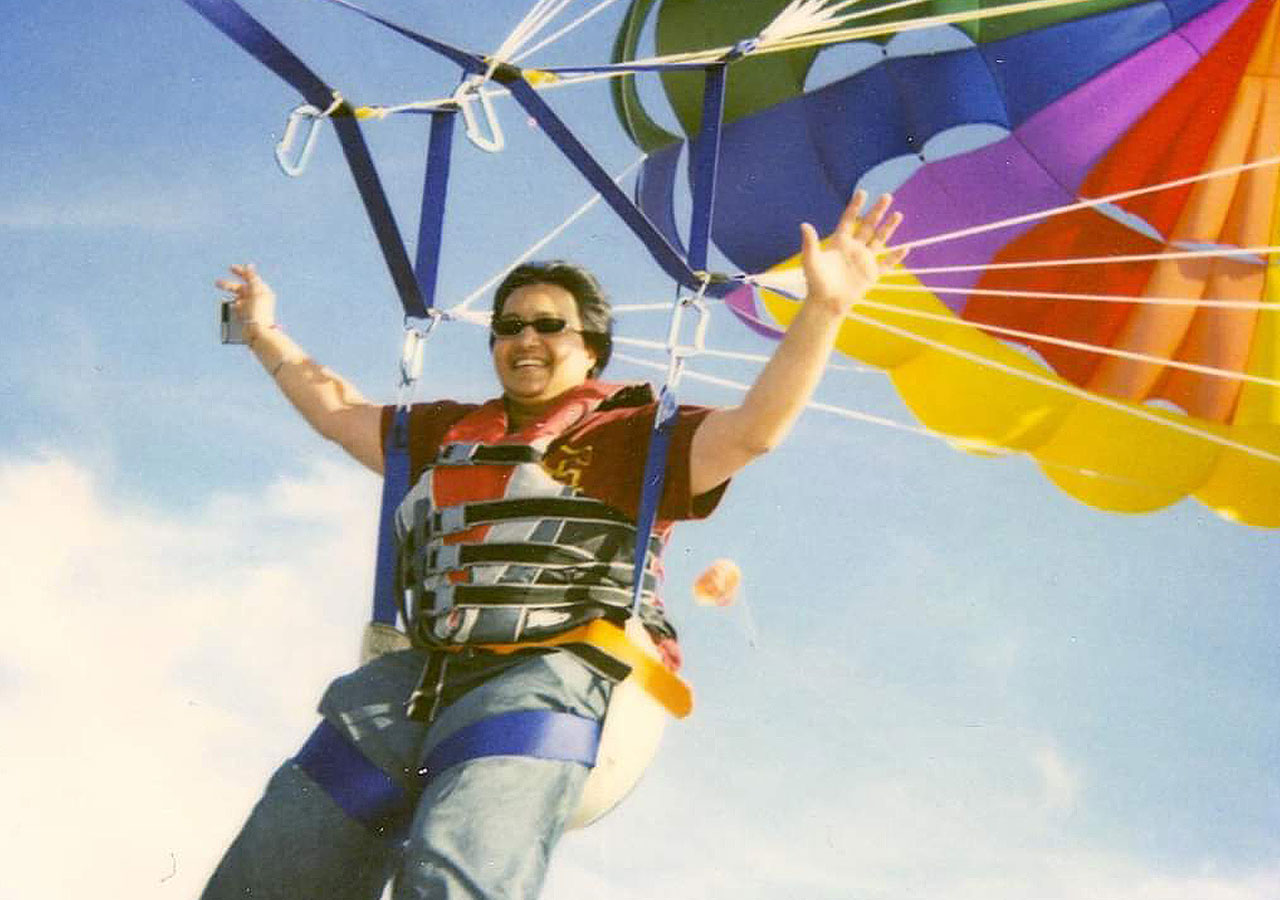  Ketty Avashia is wearing jeans, a T-shirt, and sunglasses and is parasailing with a rainbow-colored chute.