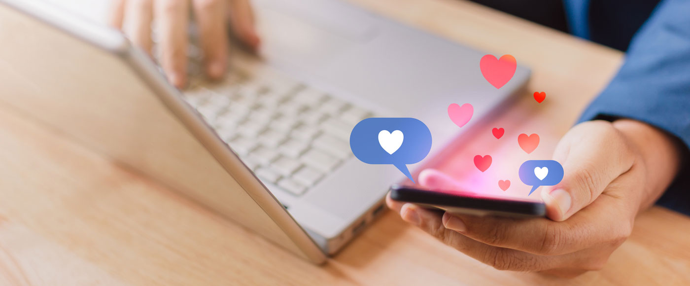 A person holds a phone with graphic heart bubbles appearing above it. The person's other hand rests on a laptop keyboard.