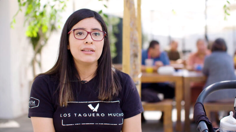 A lady wearing a Los Taquero Mucho T-shirt is interviewed on camera.