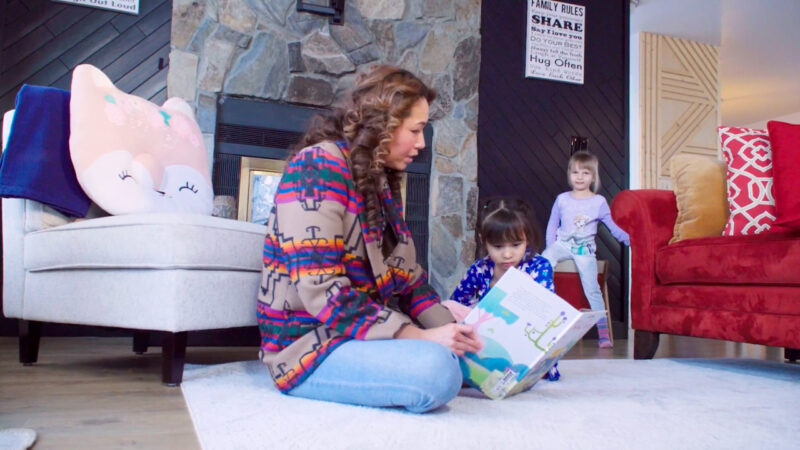A woman and a child are sitting on the family room floor reading a book, while another child sits on a chair behind them and looks on.