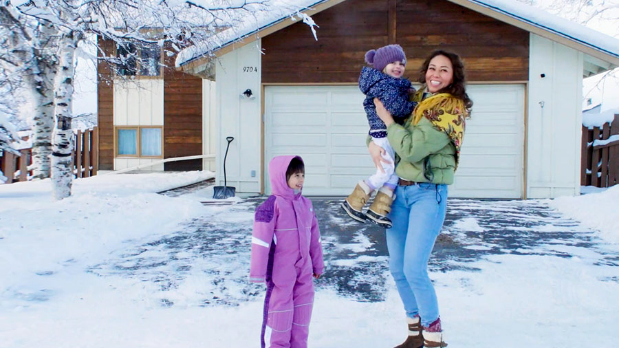 A woman stands in the snow-covered driveway of her home, holding one child in her arms while another child stands next to them.