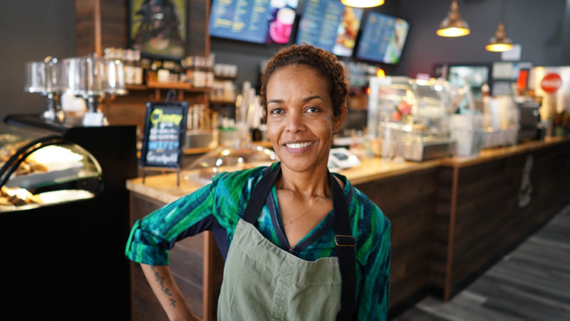 A woman in an apron stands in front of a coffeehouse counter.