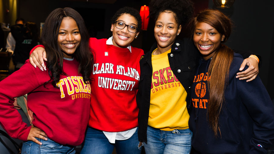 Four college students stand with their arms around each other, smiling at the camera. They are each wearing a sweatshirts from an HBCU.