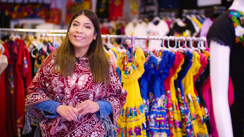 A woman wearing a brightly colored blouse stands smiling in front of a rack of colorful clothing.