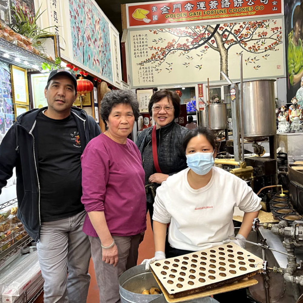 Golden Gate Fortune Cookie Company owners Kevin and Nancy Chan and an employee with BeChinatown’s Lily Lo next to a fortune cookie machine.