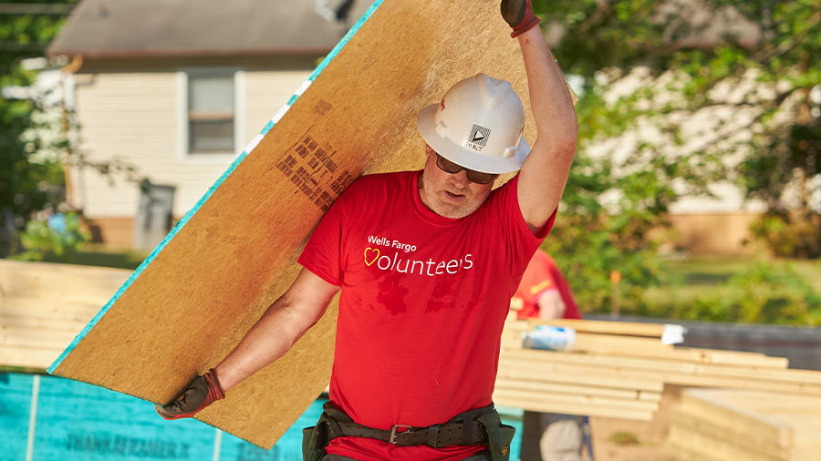 A person in a hardhat carries a plywood panel at a construction site.