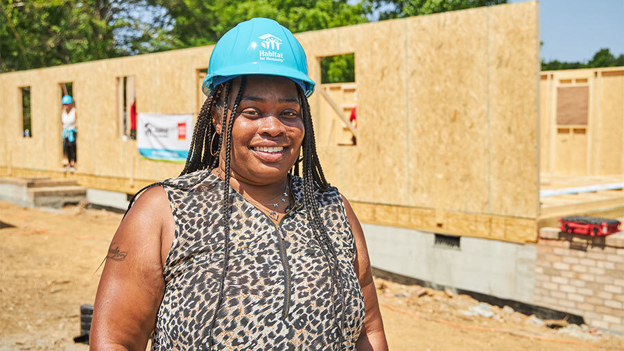 A woman in a hardhat smiles as she stands in front of a partially built home.