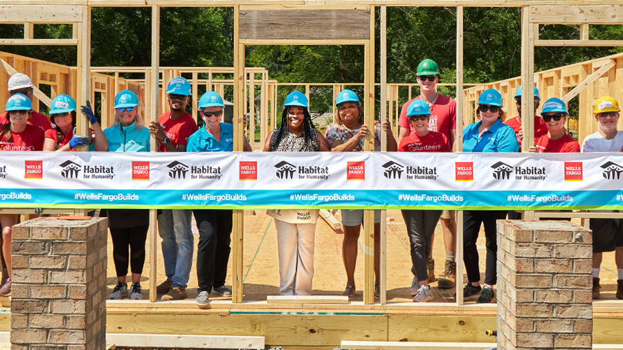 A group of people in hardhats stand inside the frame of a partially built home. A Wells Fargo and Habitat for Humanity banner is in front of them.