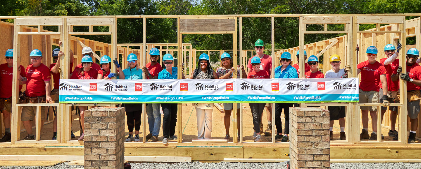 A group of people in hardhats stand inside the frame of a partially built home. A Wells Fargo and Habitat for Humanity banner is in front of them.