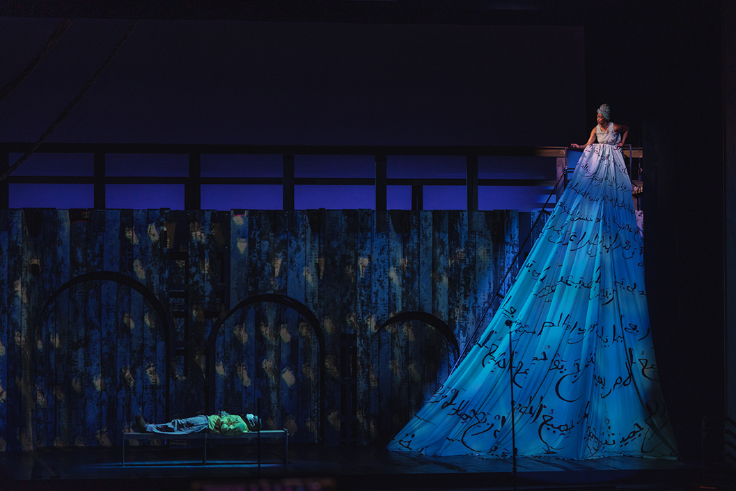  On a stage, a man is lying down while a woman is standing about 10 feet off the ground wearing a large dress with Arabic writing on it.