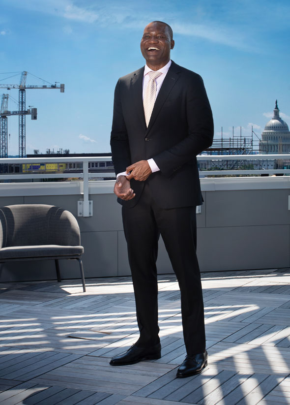 A man in a dark suit stands on a rooftop smiling at the camera.