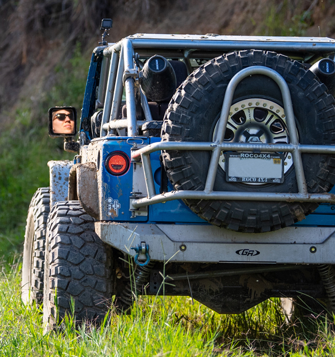 A blue Jeep sits at the base of a mountain. A person’s face is visible in the driver’s side mirror.