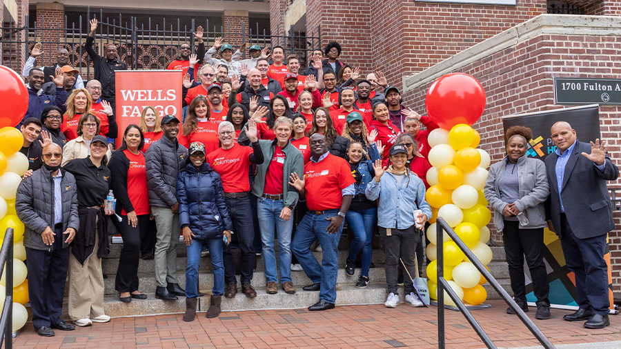 A group of volunteers standing waving on steps near a Wells Fargo banner. Red, white, and gold balloons are on either side.