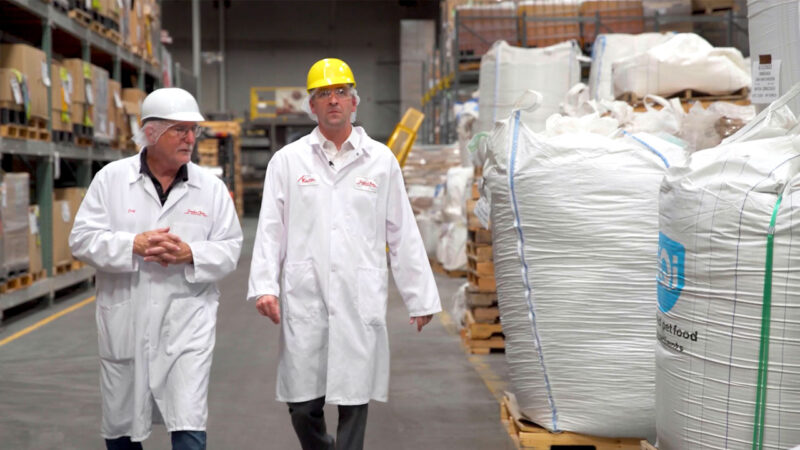 Two men in hard hats and white lab coats are walking in a factory surrounded by large sacks of food materials.