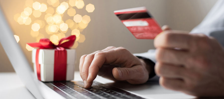 A close up of a person's hands. One hand is typing on a laptop and the other is holding a red credit card. There is a gift box on the table with the laptop and holiday lights in the background.