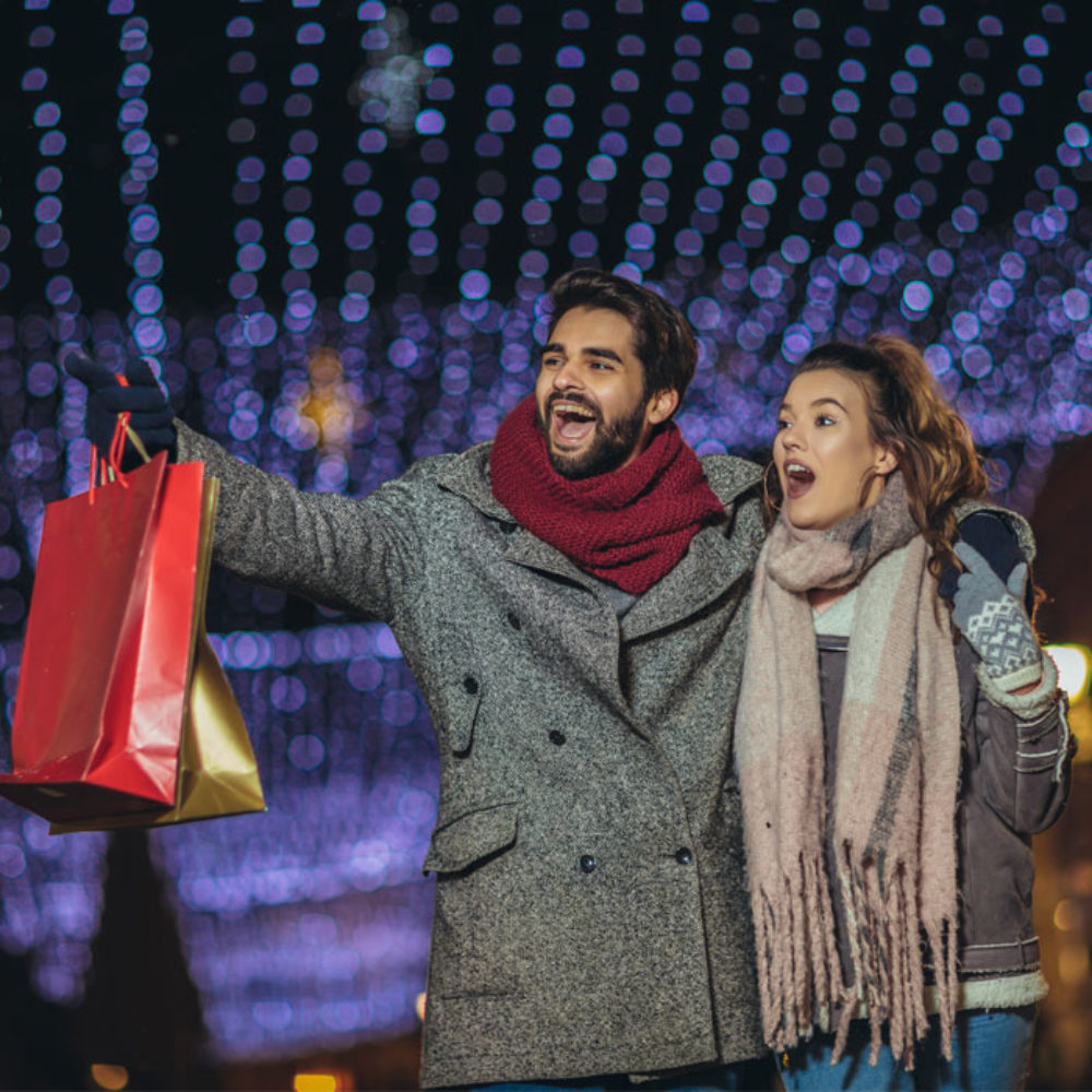 A cheerful man and woman bundled up in coats, scarfs, and gloves are standing in front of holiday lights. The man is holding gift bags and pointing at something offscreen.