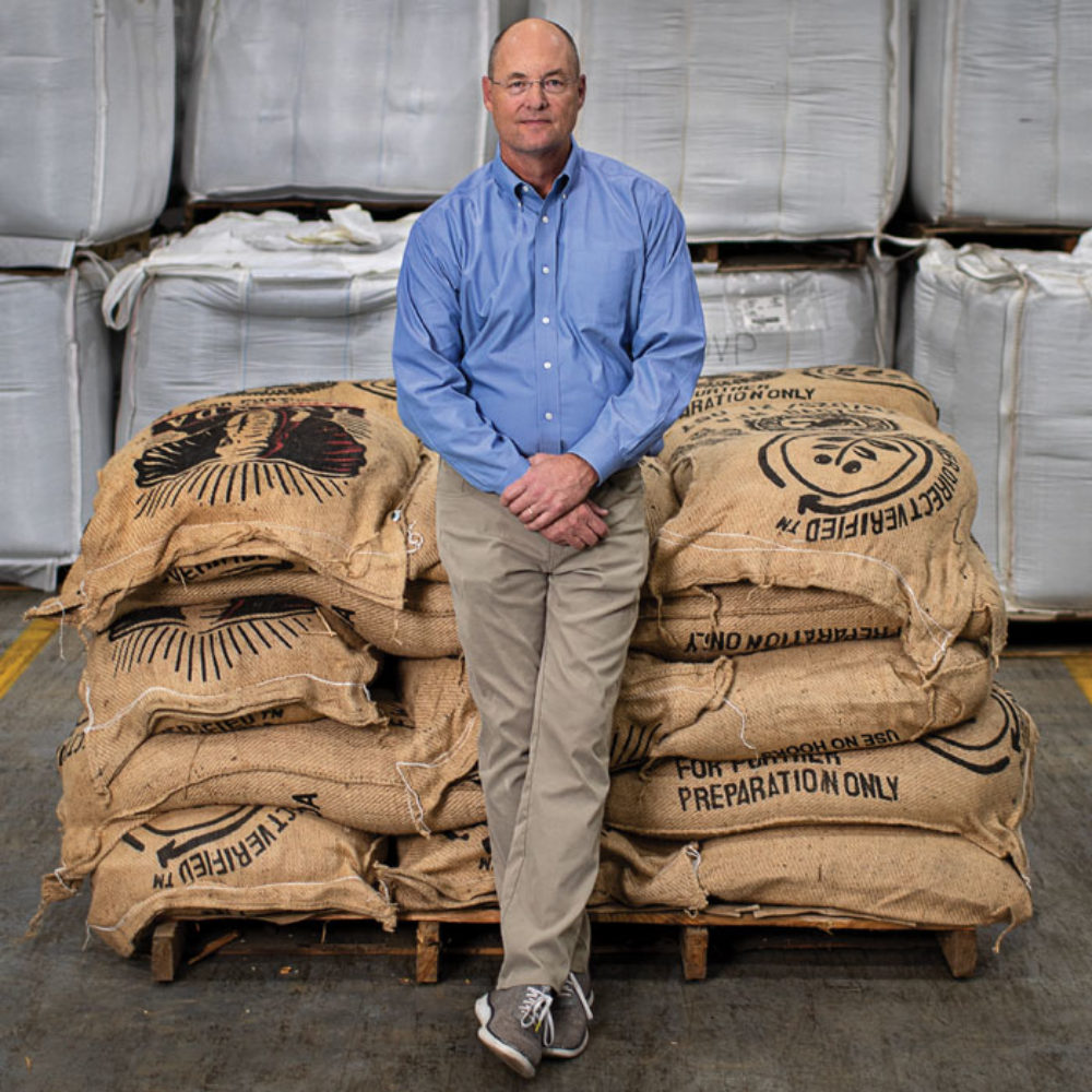 Scott Ford in a blue button-down shirt sits against burlap bags of coffee beans.