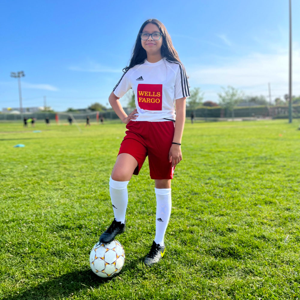 A young person in a soccer kit with a Wells Fargo logo stands on a soccer field with one foot on top of a soccer ball.