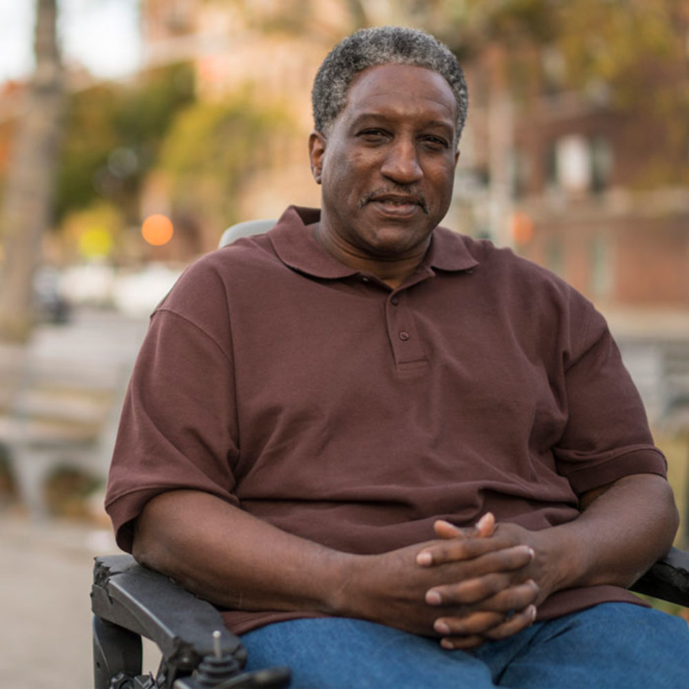 A man wearing a brown shirt and jeans is sitting in a wheelchair outside.