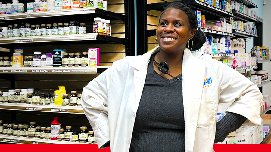Dr. TaQuina Warren stands smiling, wearing a lab coat and a headset around her neck, inside her pharmacy.