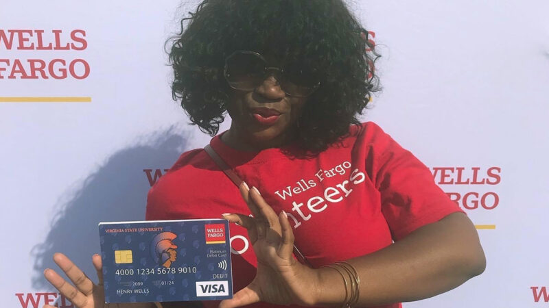 Shemesha Burton stands in front of a white background that says Wells Fargo wearing a red Wells Fargo volunteers T-shirt and holding an oversized debit card.