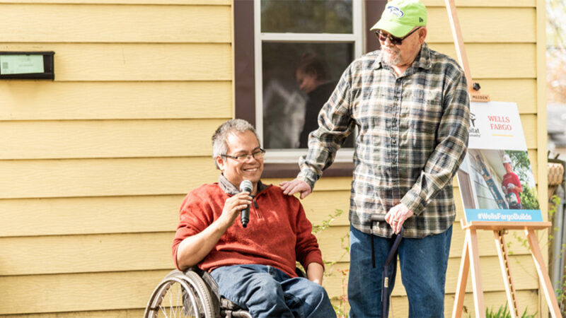A man in a wheelchair sits in front of a house and holds a microphone. A man with a cane stands next to him smiling.
