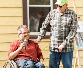 A man in a wheelchair sits in front of a house and holds a microphone. A man with a cane stands next to him smiling.