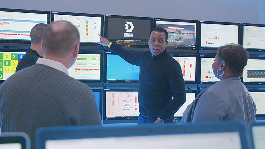 A group of people stand around a wall of tv screens while a man points at information on a screen.
